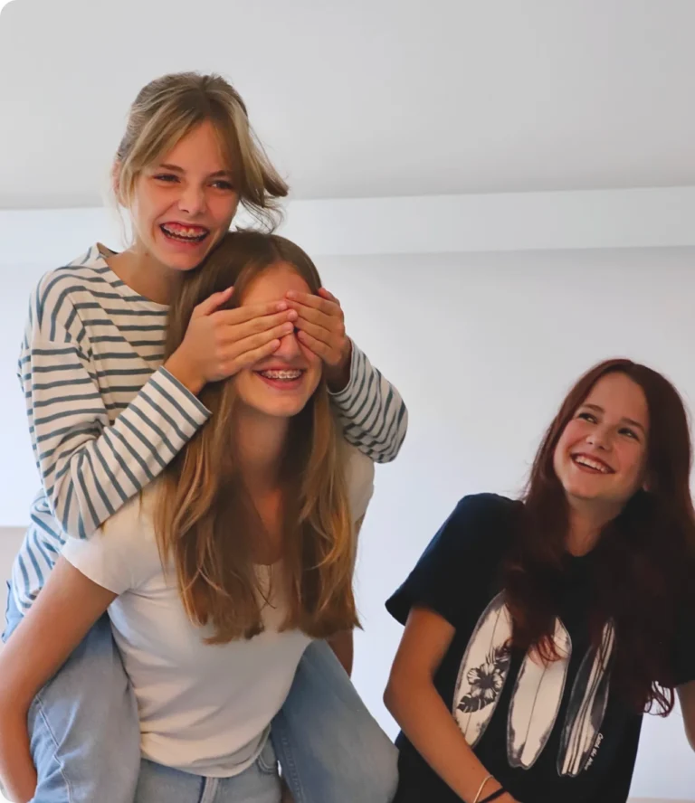 Drei lachende Jugendliche, ein Mädchen hält einem anderen spielerisch die Augen zu