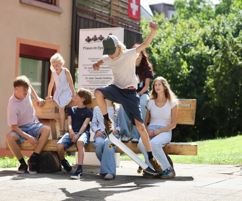 Jugendliche auf der Holzbank vor dem Eingang der Praxis im Spatzenhof, ein Junge springt mit dem Skateboard
